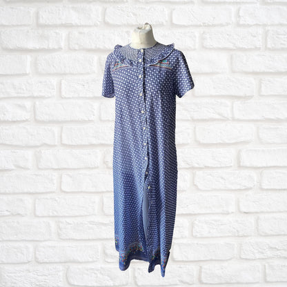 A vintage 70s blue and white geometric print dressing gown with multicolored florals, featuring short sleeves, bodice smocking, and a frill detail, displayed on a mannequin against a white brick wall.