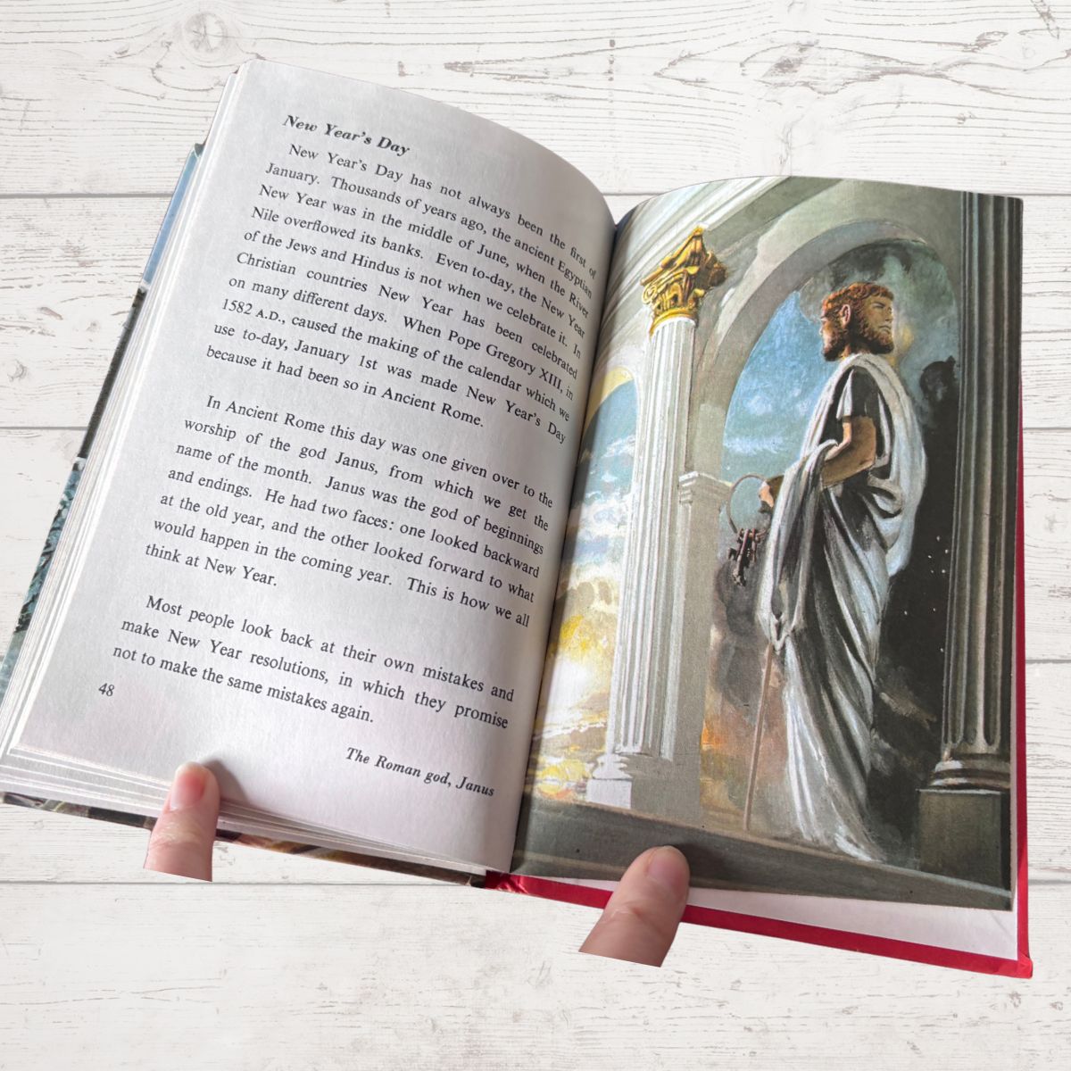 An open hardcover book being held by two hands, showing printed text on the left page and a colorful illustration of a robed figure in an architectural scene on the right page. The book lies on a light whitewashed wood surface.