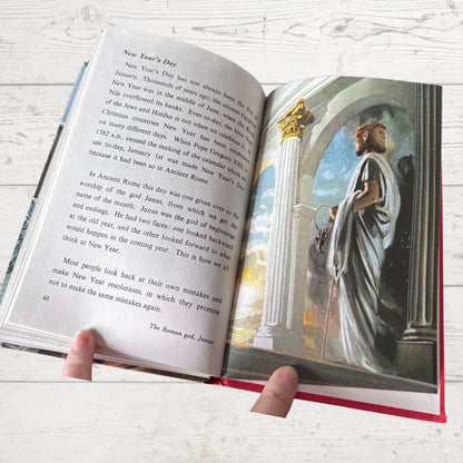 An open hardcover book being held by two hands, showing printed text on the left page and a colorful illustration of a robed figure in an architectural scene on the right page. The book lies on a light whitewashed wood surface.