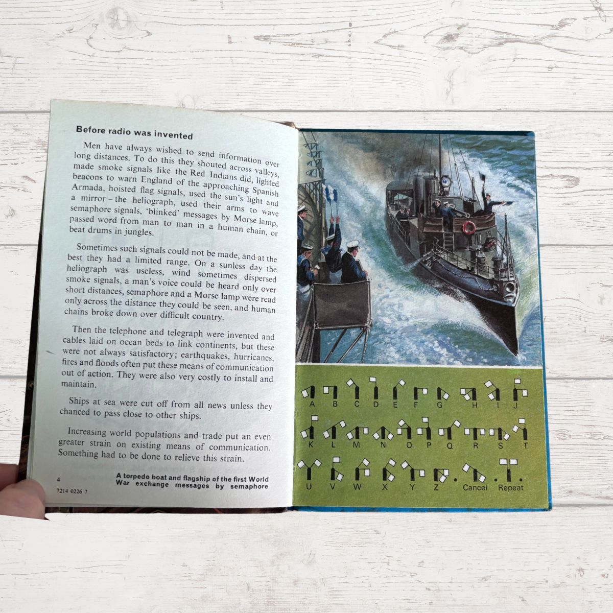 A hand holds a vintage paper booklet or book page showing printed text on the left and a colorful illustration of a ship on the right, placed on a light wood surface.