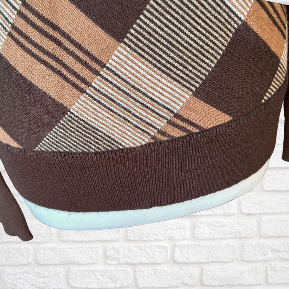 Close-up of a knit sweater with a brown, beige, and white plaid pattern, featuring a ribbed brown cuff and a light blue inner lining, set against a white brick wall background.