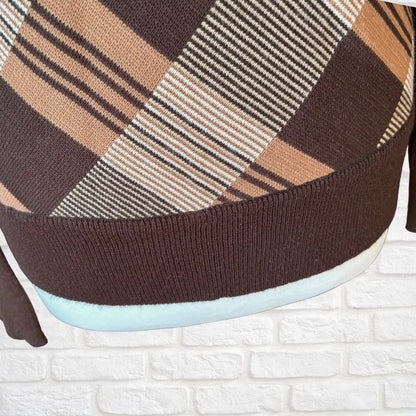 Close-up of a knit sweater with a brown, beige, and white plaid pattern, featuring a ribbed brown cuff and a light blue inner lining, set against a white brick wall background.
