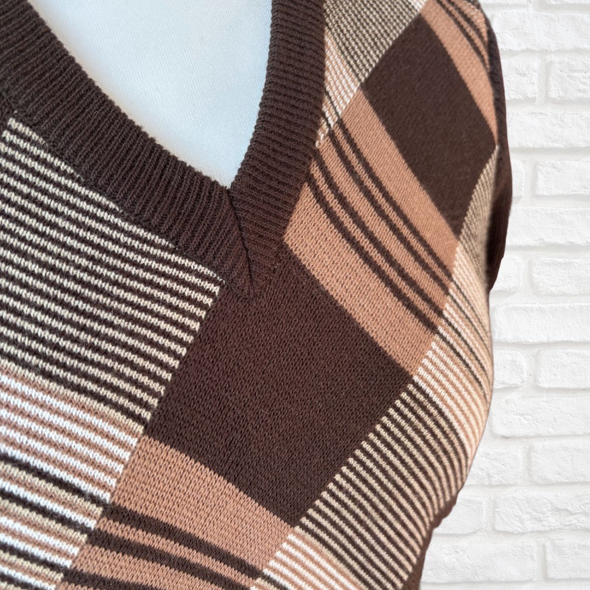 A close-up of a knit V-neck sweater on a mannequin, featuring a geometric plaid pattern in dark brown, tan, beige, and white, set against a white brick wall background.