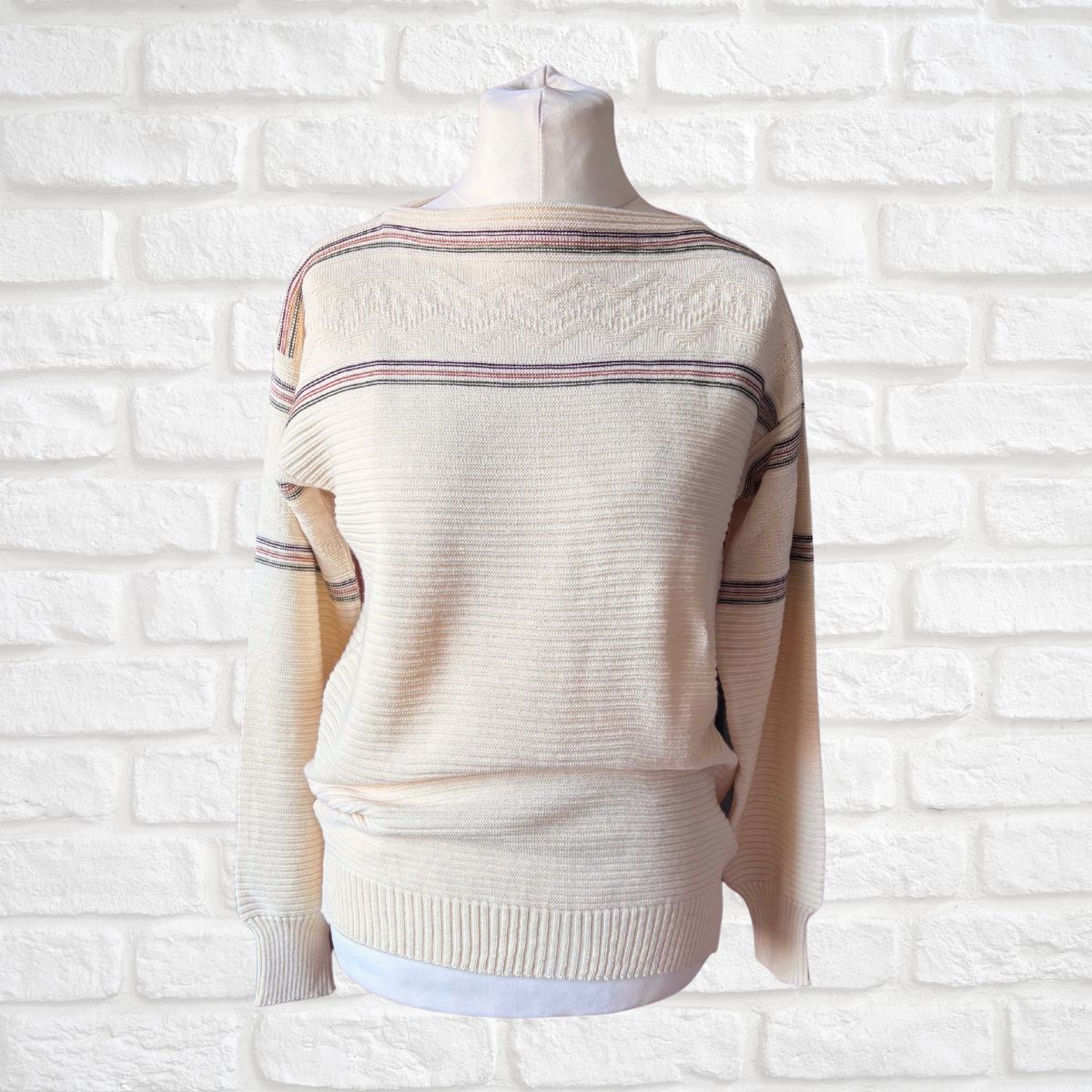 A cream-colored knit sweater with thin horizontal stripes in green and brown , displayed on a mannequin torso against a white brick wall background.