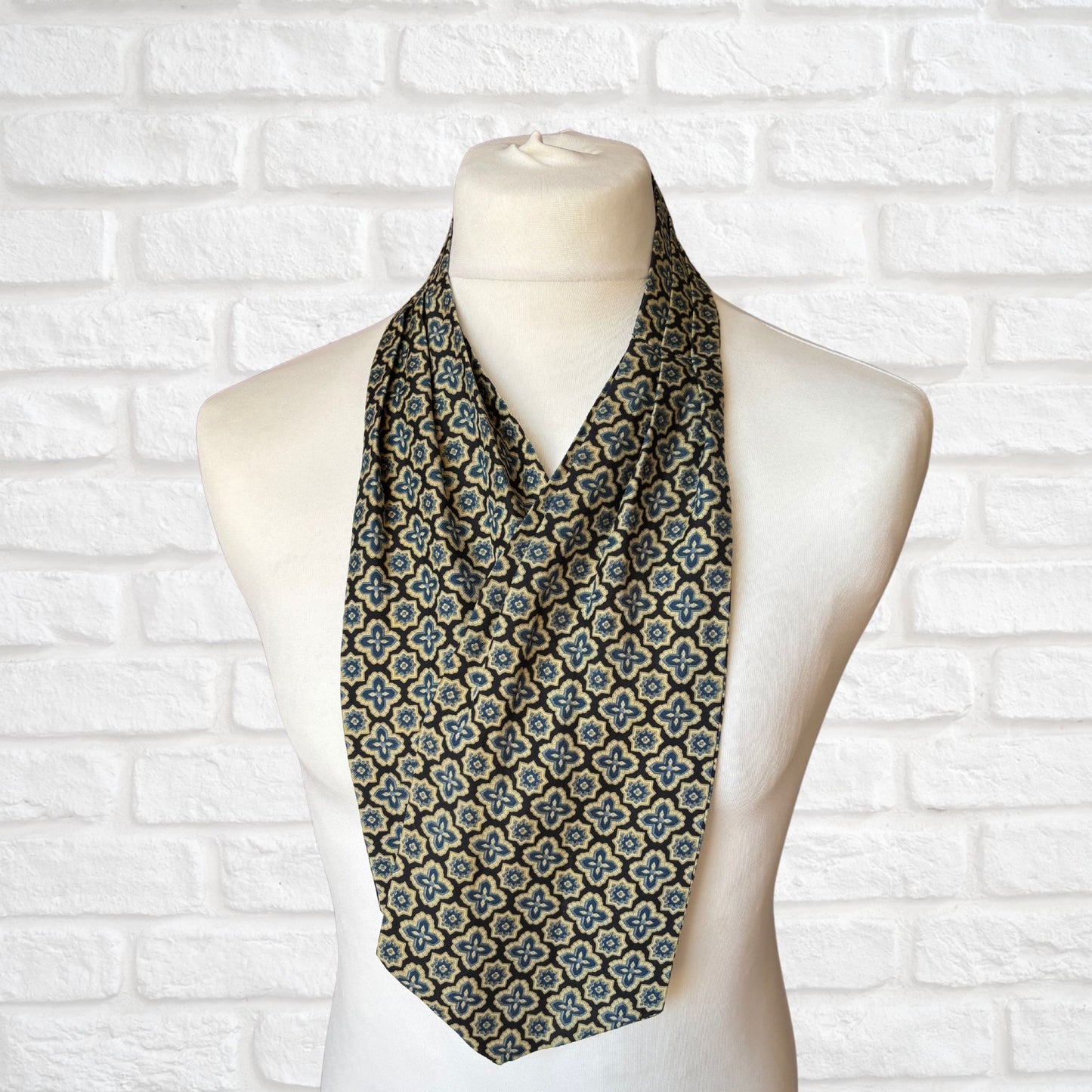 A vintage Austin Reed cravat featuring a black necktie with a cream, gold, and blue stylized floral print, displayed on a mannequin against a white brick wall background.