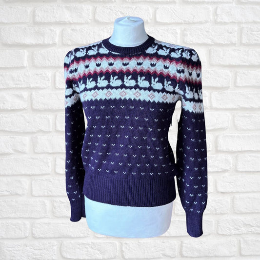 A dark blue knit sweater with a white and red Nordic fair isle pattern across the chest and sleeves, displayed on a white mannequin torso against a white brick wall background.