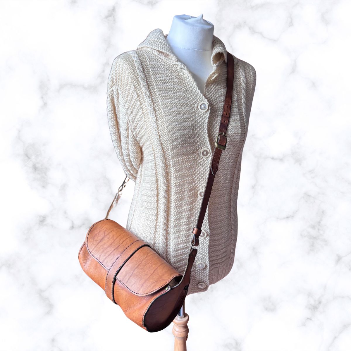 A tan leather crossbody bag with a brown shoulder strap, displayed on a cream-colored knit sweater dress mannequin against a white marble background.