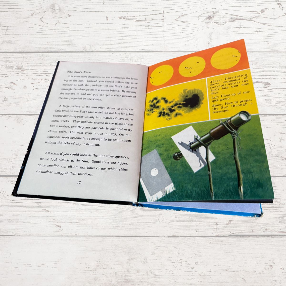 An open children's book titled 'The Sun's Place' laid flat on a white wood surface. The left page contains black text, and the right page features a colorful illustration with yellow sun circles, a black-and-white solar eclipse, and a telescope on a green landscape.