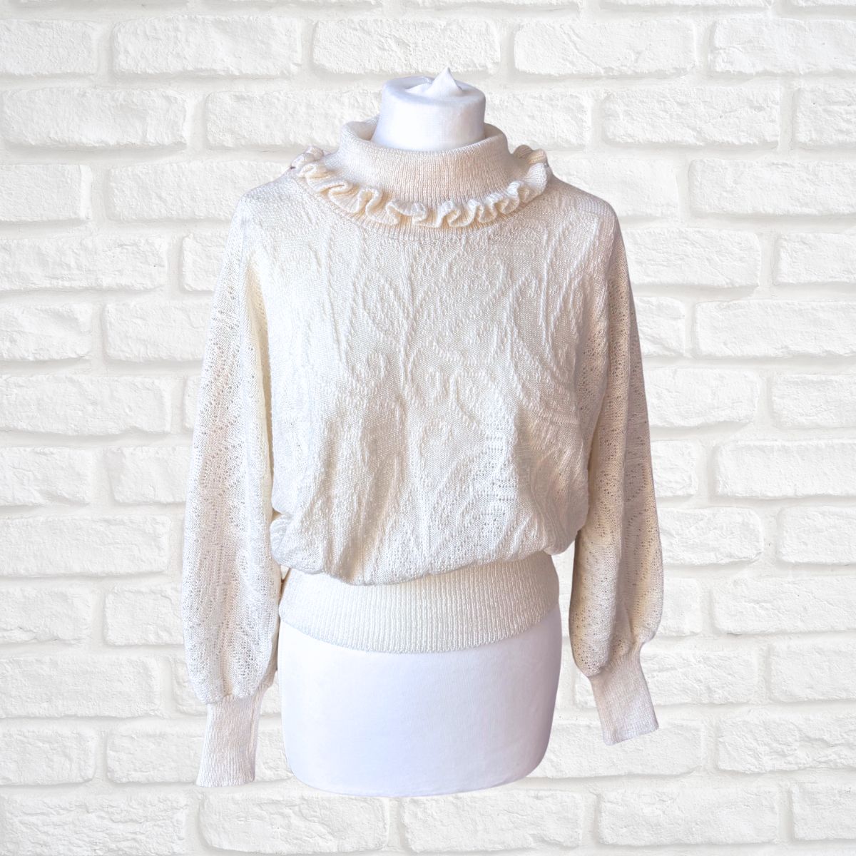 An off-white textured knit sweater with a ruffled mock neck, displayed on a white mannequin torso against a white brick wall background.