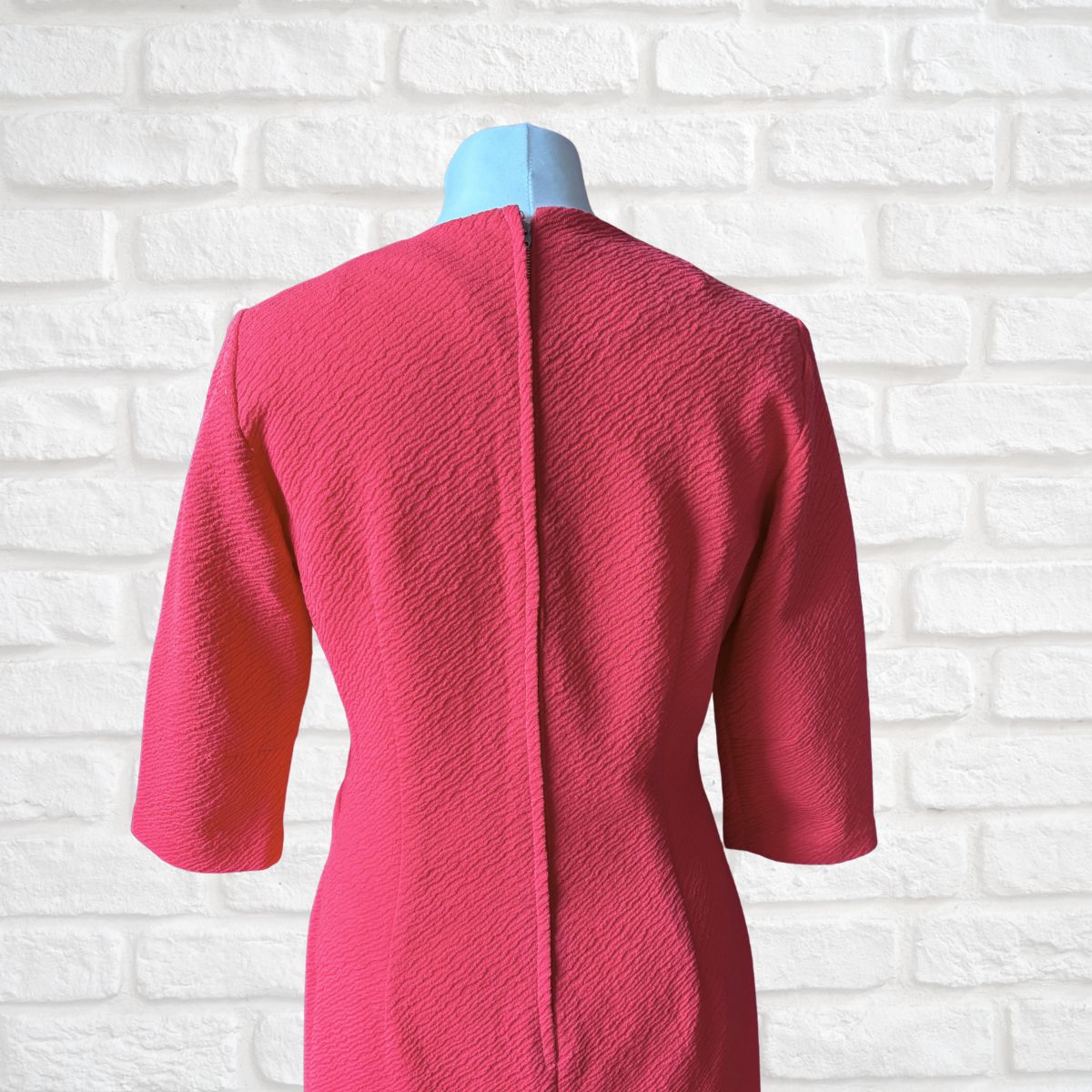 A bright pink textured dress with three-quarter sleeves, displayed on a light blue mannequin torso against a white brick wall background.