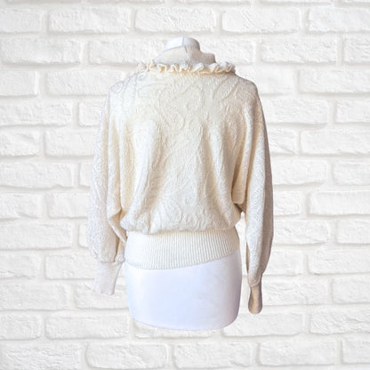 An off-white textured knit sweater with a ruffled mock neck and lace sleeves, displayed on a white mannequin against a white brick wall background.