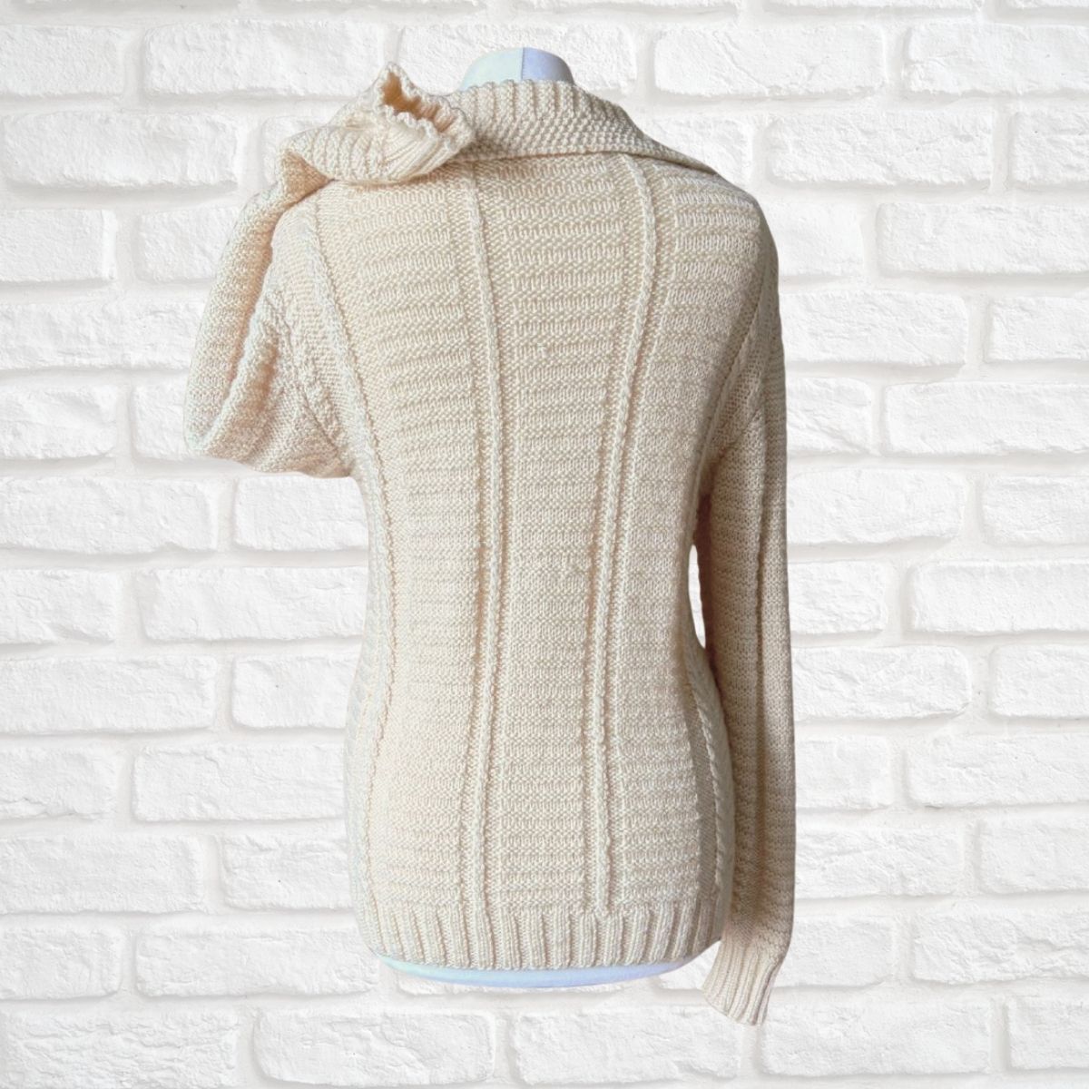 A cream-colored cable-knit sweater with a high ruffled mock neck, displayed on a mannequin torso against a white brick wall background.