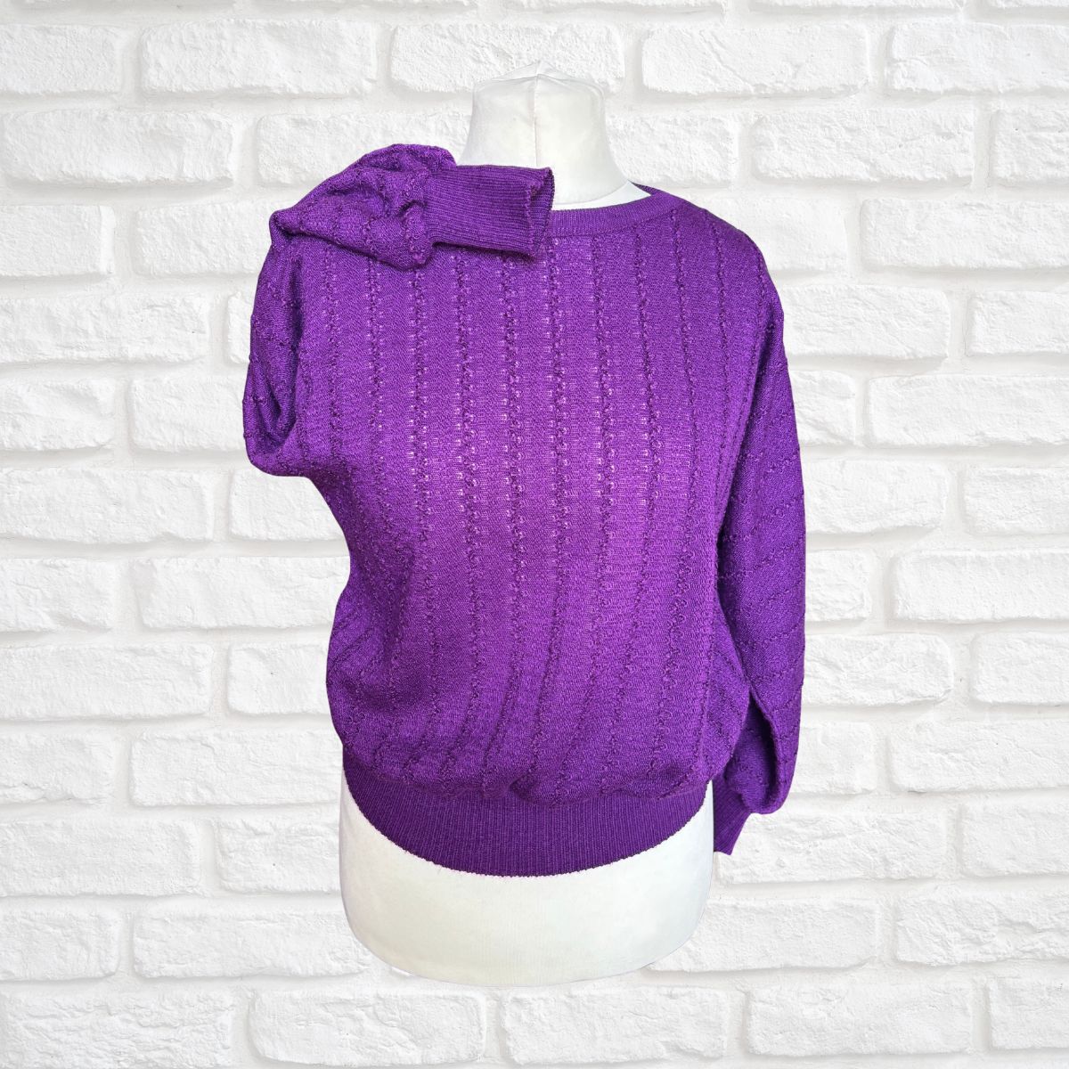 A purple cable-knit sweater displayed on a white mannequin torso against a white brick wall background.