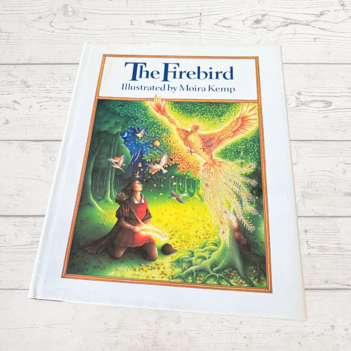 The Firebird illustrated book by Moira Kemp, featuring a white hardcover and a colorful cover illustration of a woman in a forest meeting a glowing bird, placed on a white wood plank surface.