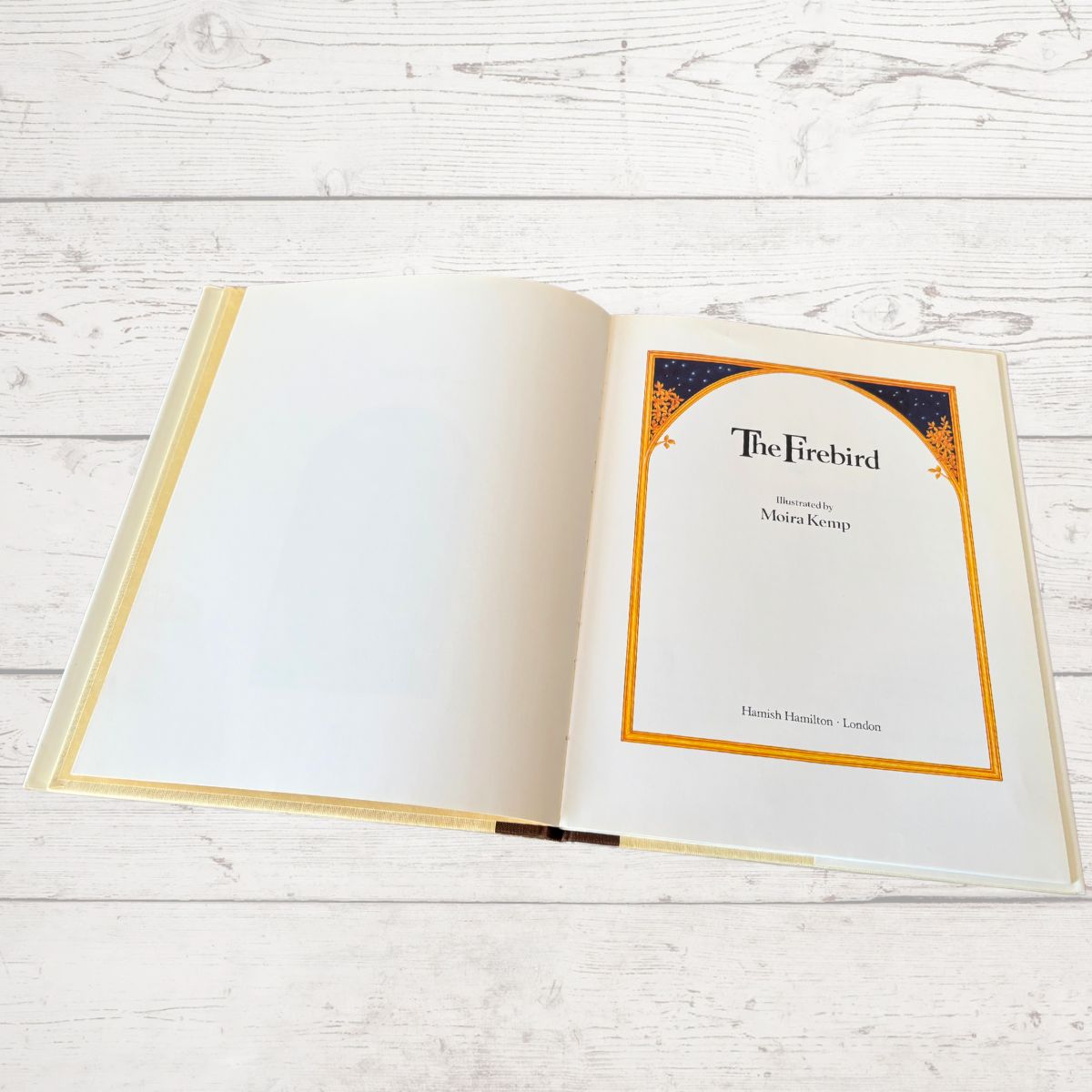 An open hardcover book titled 'The Firebird' with illustrated pages, photographed on a light whitewashed wood plank surface.