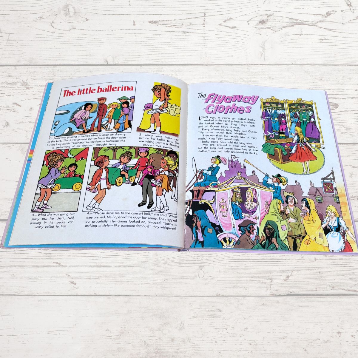 An open children's book spread laid on a white wood surface, showing two illustrated pages with colorful cartoon scenes and text including the titles 'The little ballerina' and 'The Fairy Godmother'.