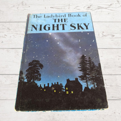 Vintage hardcover book titled 'The Ladybird Book of The Night Sky' featuring a night sky illustration with stars, silhouettes of trees and houses, photographed on a white wood-plank surface.