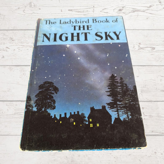 Vintage hardcover book titled 'The Ladybird Book of The Night Sky' featuring a night sky illustration with stars, silhouettes of trees and houses, photographed on a white wood-plank surface.