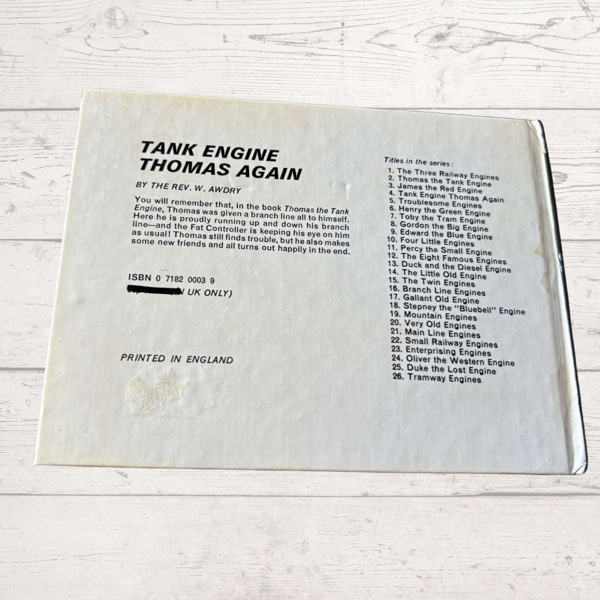 Back cover of a book titled 'Tank Engine Thomas Again' with text and ISBN information on a light wooden surface.