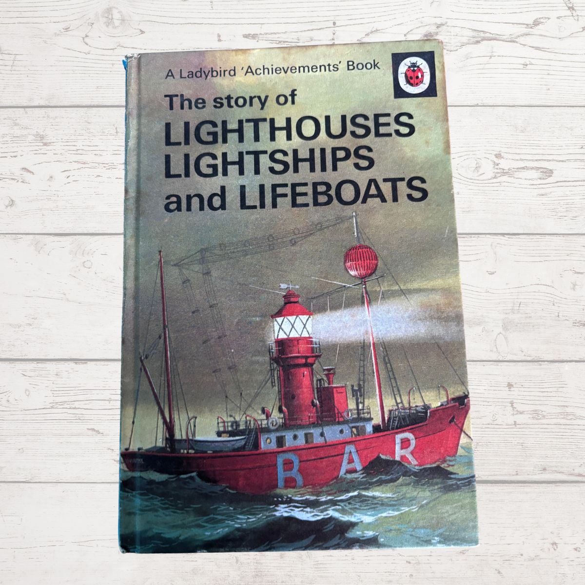 Vintage hardcover book titled 'The story of Lighthouses Lightships and Lifeboats' by Ladybird Achievements, featuring a red lifeboat illustration on the cover, placed on a light wood background.