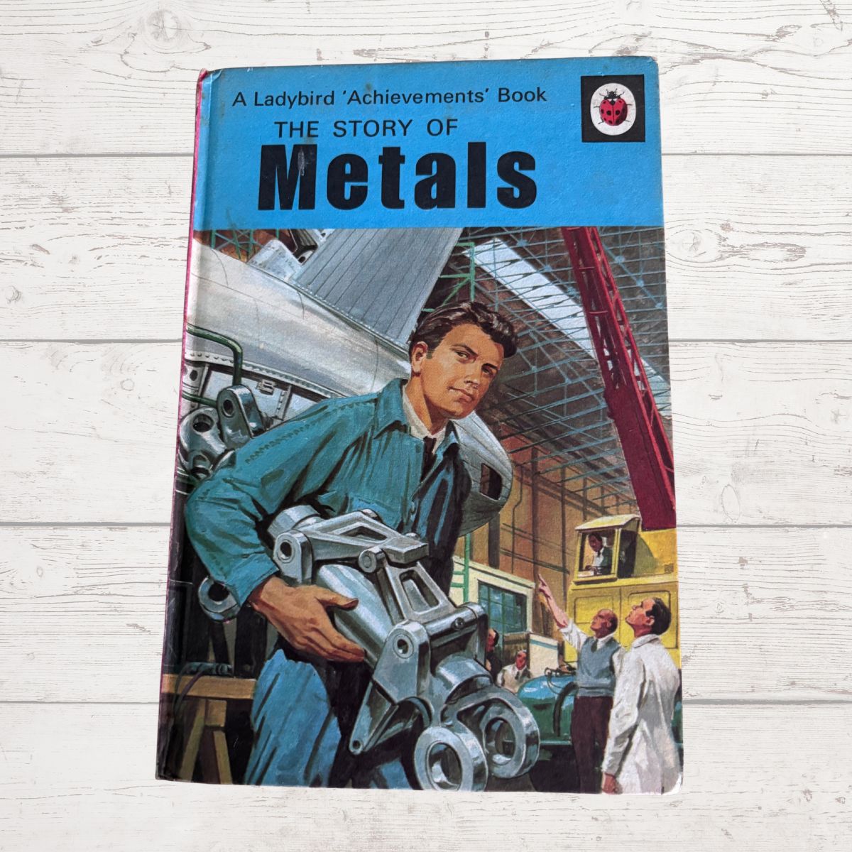 Vintage  book titled 'The Story of Metals' from the Ladybird Achievements series, featuring a colorful illustration of a young mechanic holding a metal engine part on a light wood plank background.