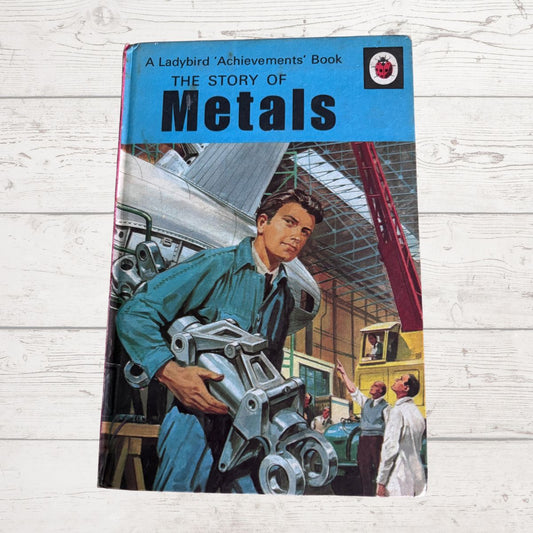 Vintage  book titled 'The Story of Metals' from the Ladybird Achievements series, featuring a colorful illustration of a young mechanic holding a metal engine part on a light wood plank background.