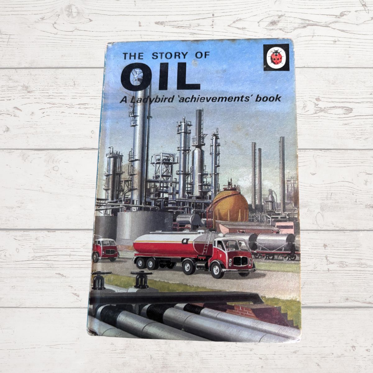 Vintage paperback book titled 'The Story of Oil' resting on a white wood plank surface. The cover features an illustration of an oil refinery with tall towers, a red tanker truck, and large silver pipelines in the foreground.
