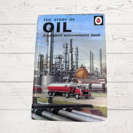 Vintage paperback book titled 'The Story of Oil' resting on a white wood plank surface. The cover features an illustration of an oil refinery with tall towers, a red tanker truck, and large silver pipelines in the foreground.