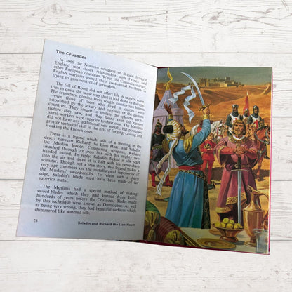 An open children's history book spread showing a page with text titled 'The Crusaders' and a colorful illustration of medieval soldiers on a desert battlefield, placed on a white wood surface.