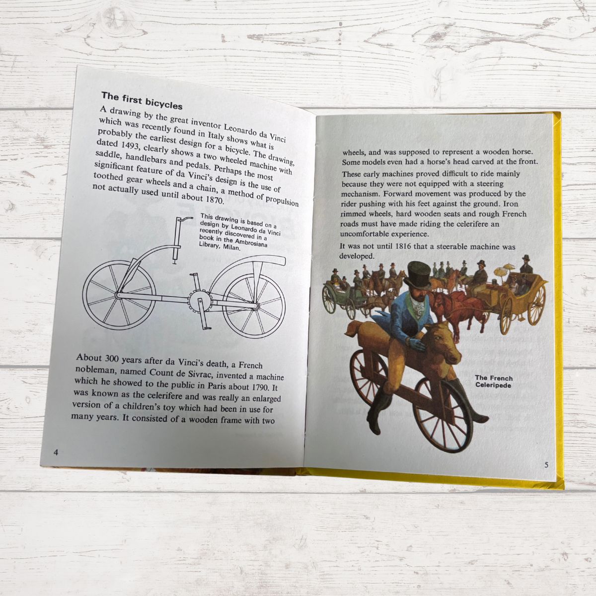 An open children's history book spread showing illustrations and text about early bicycles and the French carriage. The pages are photographed on a white wood surface.