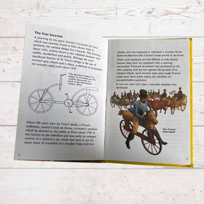 An open children's history book spread showing illustrations and text about early bicycles and the French carriage. The pages are photographed on a white wood surface.