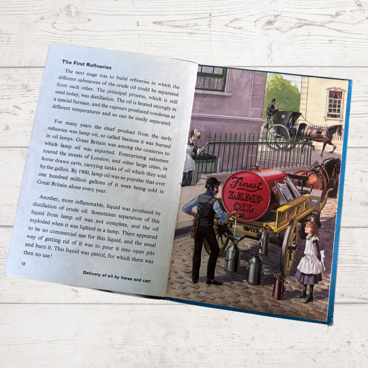 An open book showing a page about the first refinery with printed text on the left and a vintage illustration on the right, photographed on a light wood surface.