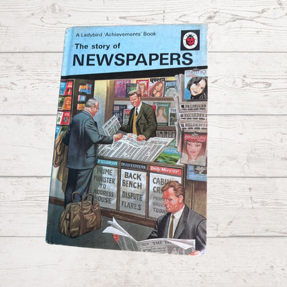 A vintage paperback book titled 'The story of NEWSPAPERS' by Ladybird Achievements, featuring a colorful illustration of a newspaper stand with people reading and selling papers, photographed on a light wood background.