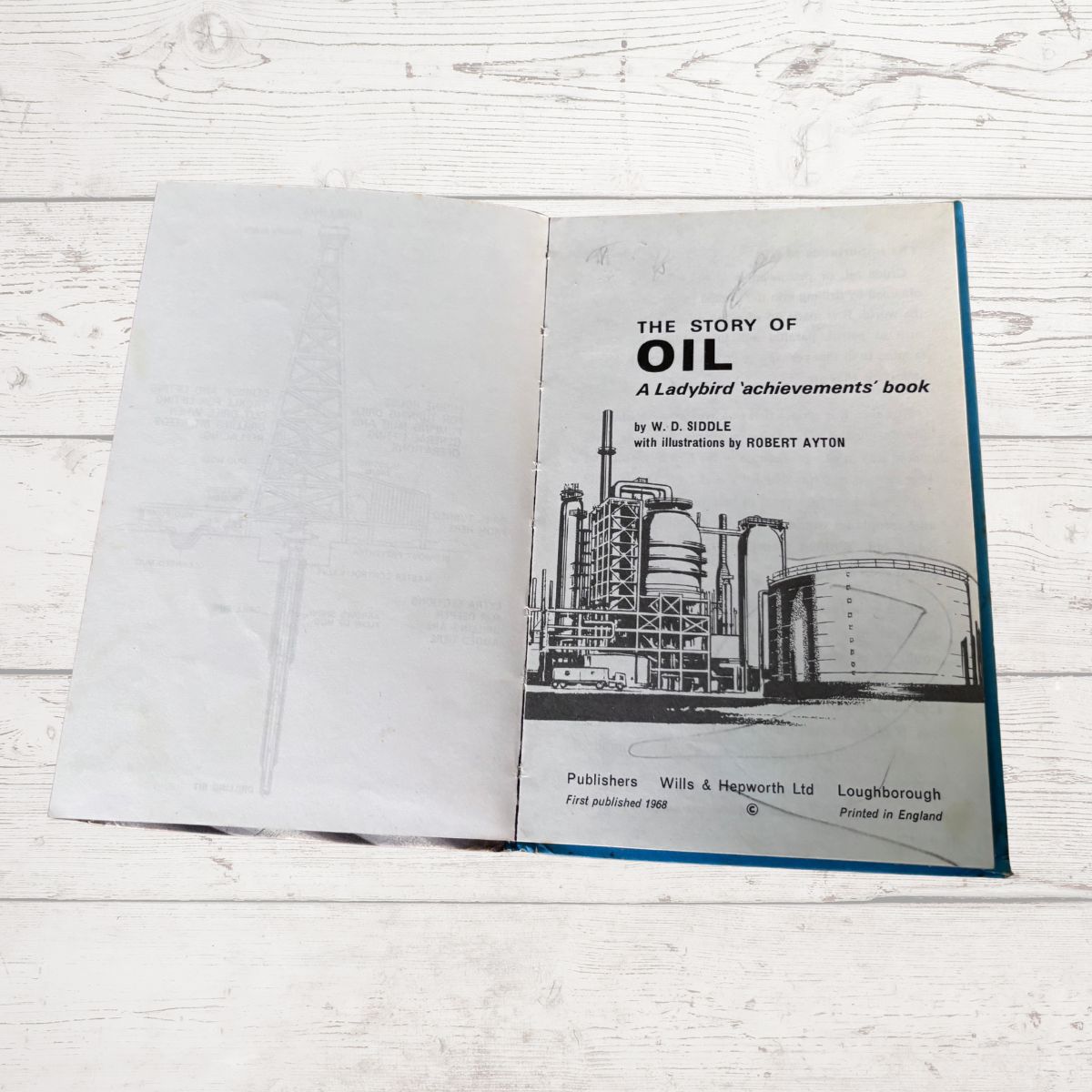 An open vintage book titled 'The Story of Oil: A Ladybird achievements' book, showing the cover page with a black-and-white illustration of an oil refinery and industrial tanks, placed on a white wood surface.