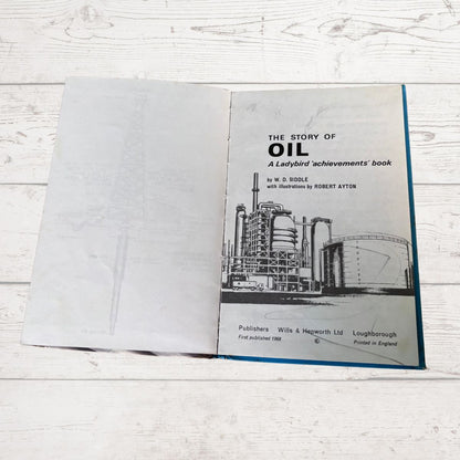 An open vintage book titled 'The Story of Oil: A Ladybird achievements' book, showing the cover page with a black-and-white illustration of an oil refinery and industrial tanks, placed on a white wood surface.