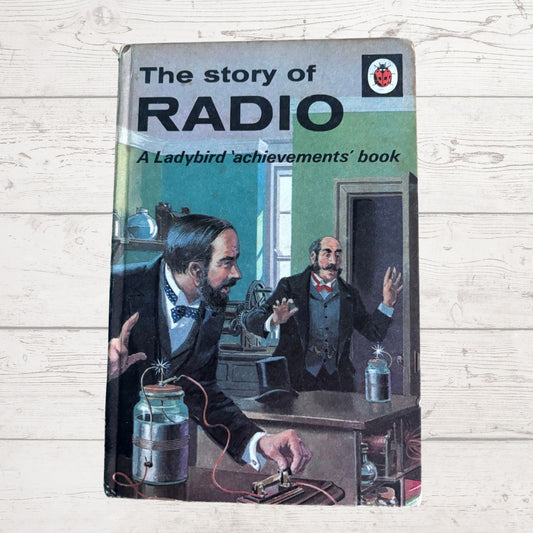 Vintage  book titled 'The story of RADIO' from the Ladybird achievements series, featuring a colorful illustration of scientists experimenting with radio on a wooden table, set against a light wood plank background.