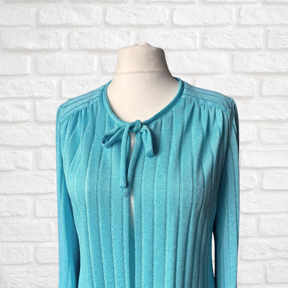 A turquoise blue ribbed knit long-sleeve knit with a tie-neck detail, displayed on a mannequin torso against a white brick wall background.