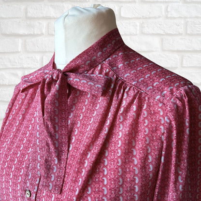 Pink and grey patterned tie neck blouse on a mannequin against a white brick wall.