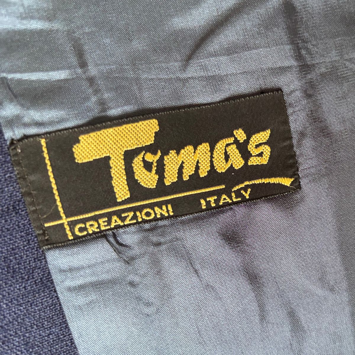 A close-up of a black fabric label with yellow embroidered text reading 'Toma's' and 'CREAZIONI ITALY' sewn onto gray clothing.