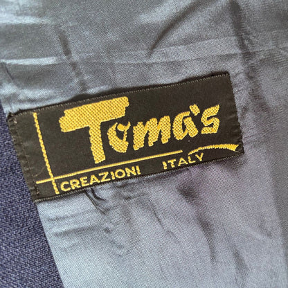 A close-up of a black fabric label with yellow embroidered text reading 'Toma's' and 'CREAZIONI ITALY' sewn onto gray clothing.