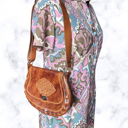 A brown leather shoulder bag with a decorative stitched patch on the front and a studded edge, shown worn crossbody over a patterned dress against a white marble background.
