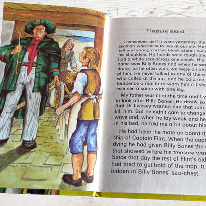 An open book showing a printed page with the heading 'Treasure Island' and a colorful illustrated scene on the left page depicting characters in a ship's cabin.