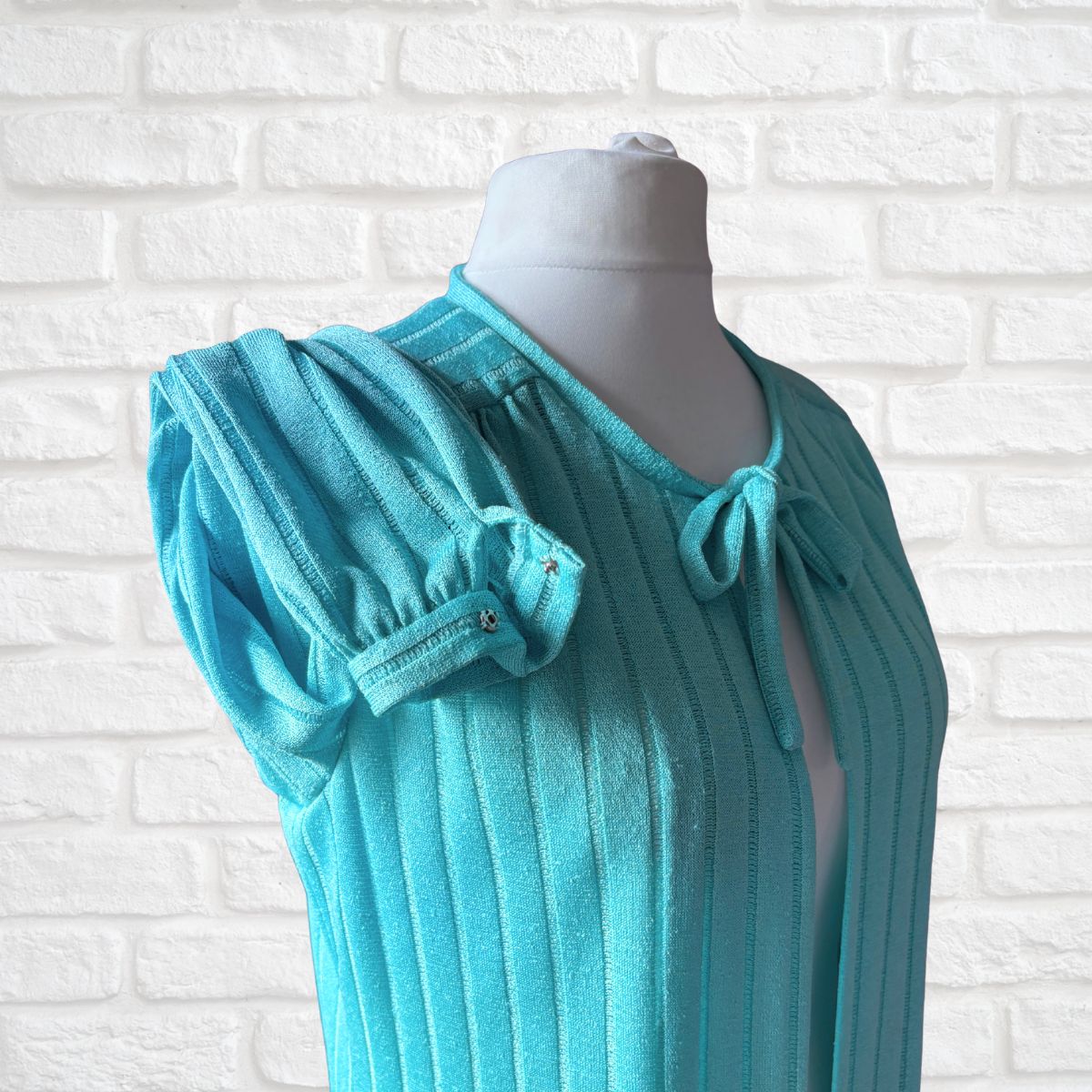 A turquoise ribbed knit housecoat with puffed short sleeves and a tie-neck detail, displayed on a white mannequin torso against a white brick wall background.