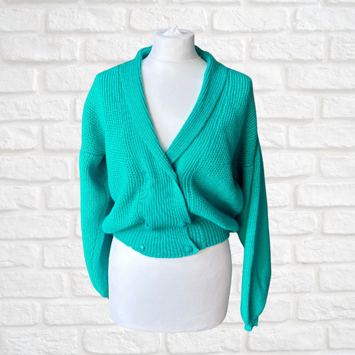 A bright turquoise knit cardigan sweater displayed on a white mannequin torso against a white painted brick wall background.