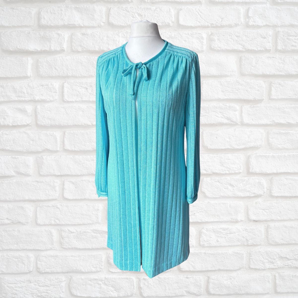 A turquoise blue ribbed knit long-sleeve cardigan with a tie-neck detail, displayed on a white mannequin torso against a white brick wall background.