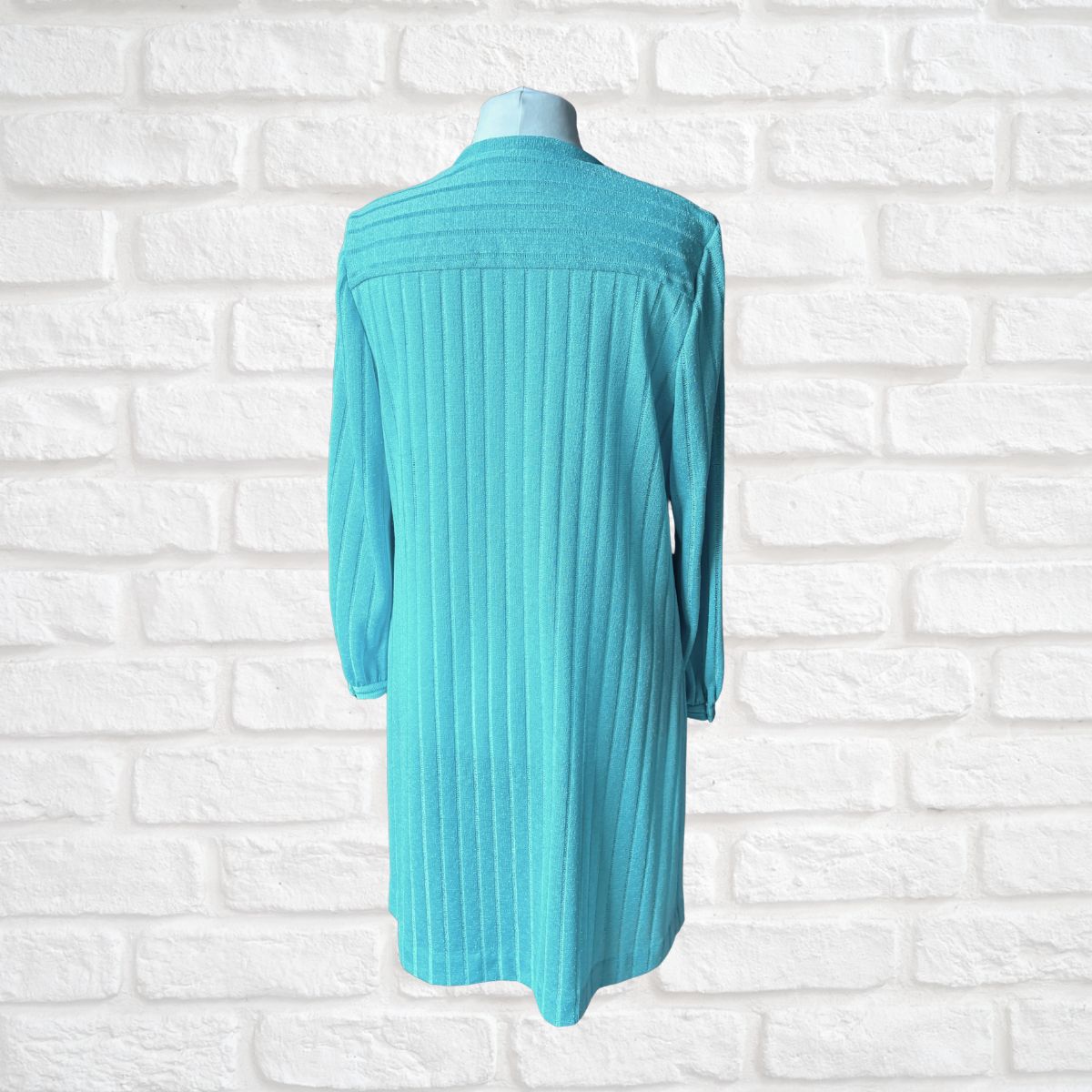 A turquoise ribbed knit long-sleeve cardigan or tunic dress displayed on a mannequin torso against a white brick wall background.