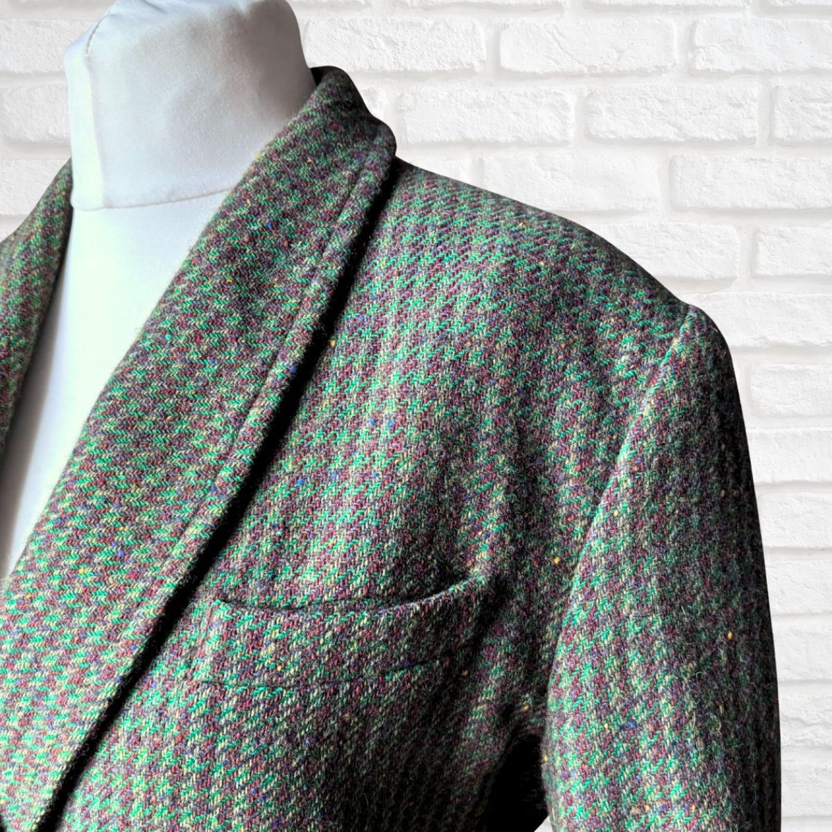 A close-up of a Womens  tweed blazer on a white mannequin torso, featuring a green and brown houndstooth pattern against a white brick wall background.