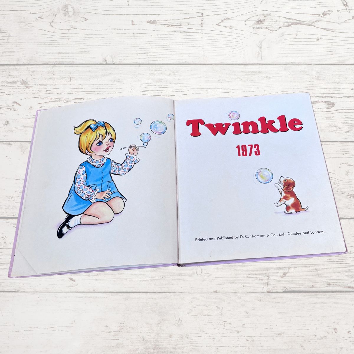 An open vintage hardcover book titled 'Twinkle 1973' laid flat on a light whitewashed wooden surface. The left page shows an illustration of a young girl in a blue dress blowing bubbles, and the right page has the title 'Twinkle 1973' in red with a small dog illustration.