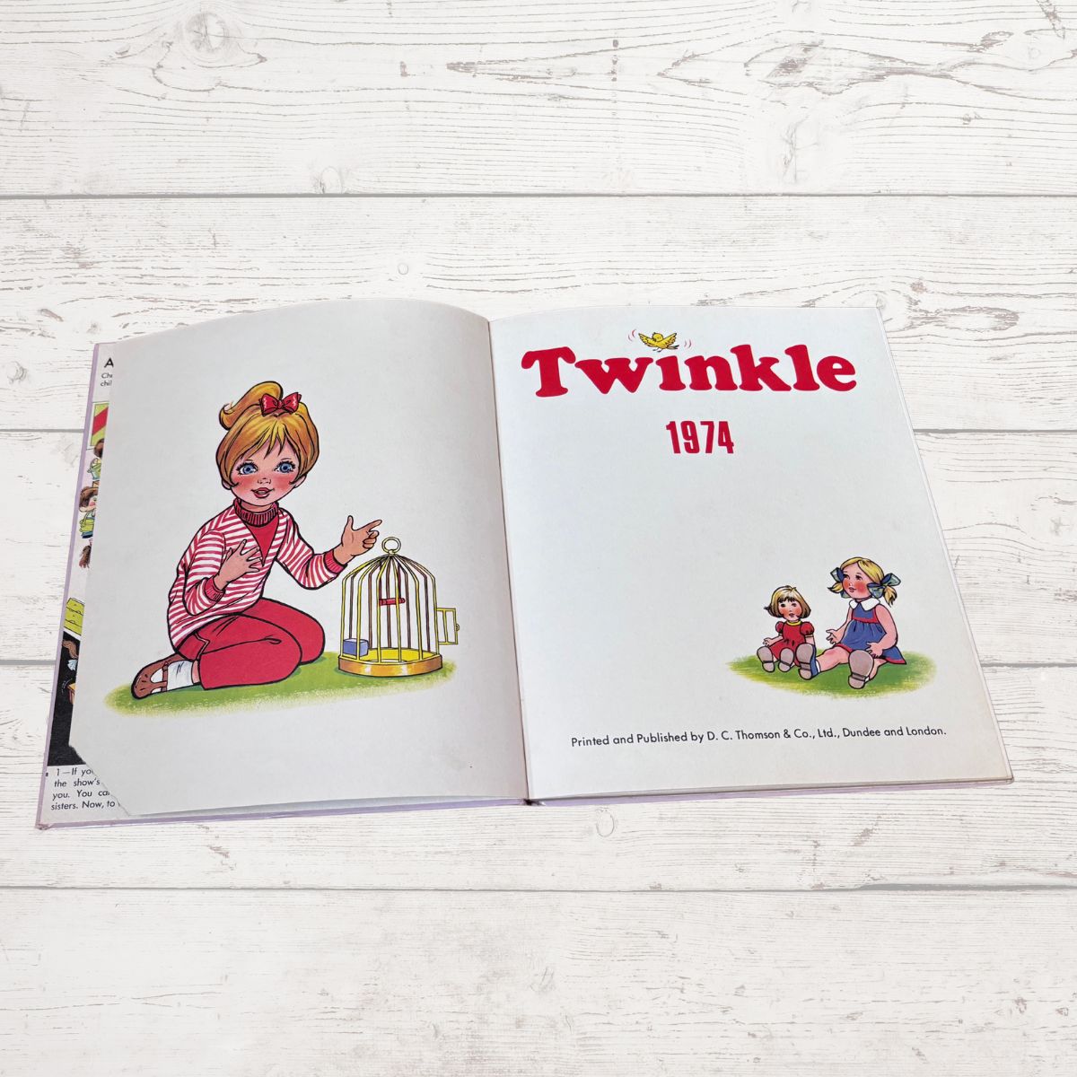 An open vintage children's book titled 'Twinkle 1974' laid flat on a white wooden surface. The left page shows an illustration of a girl in a pink striped shirt and red pants sitting beside a small birdcage, while the right page has the title 'Twinkle 1974' in red and small character illustrations.
