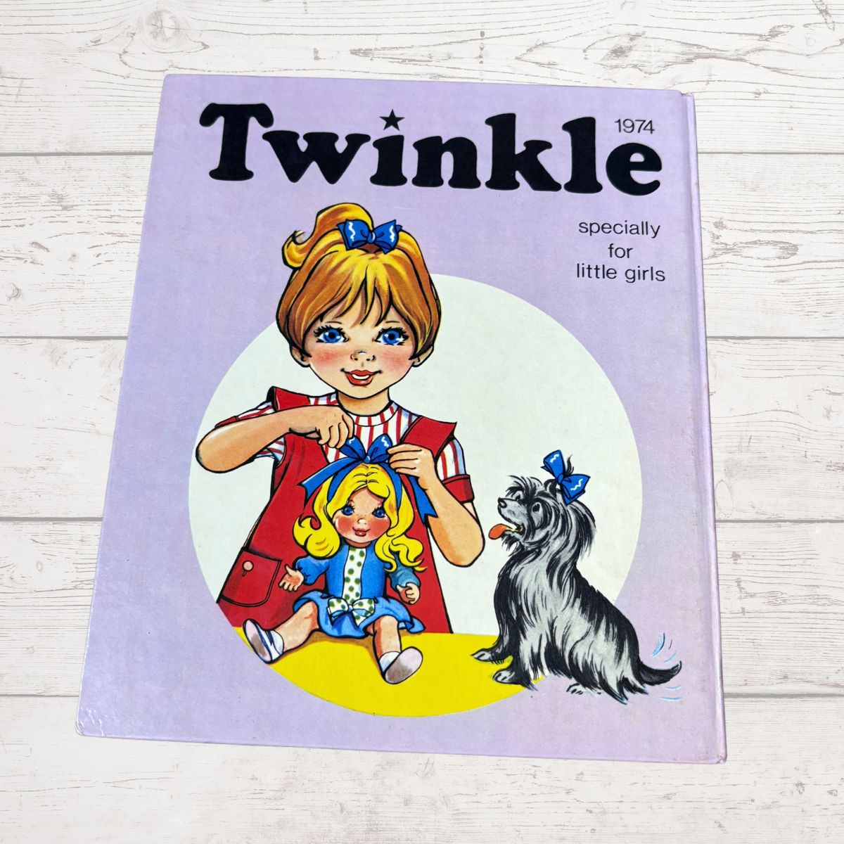 A vintage 1974 children's book or magazine titled 'Twinkle' with the subtitle 'specially for little girls,' shown lying on a whitewashed wood surface. The cover features an illustration of a girl in a red dress holding a doll, with a small black-and-white dog beside her.